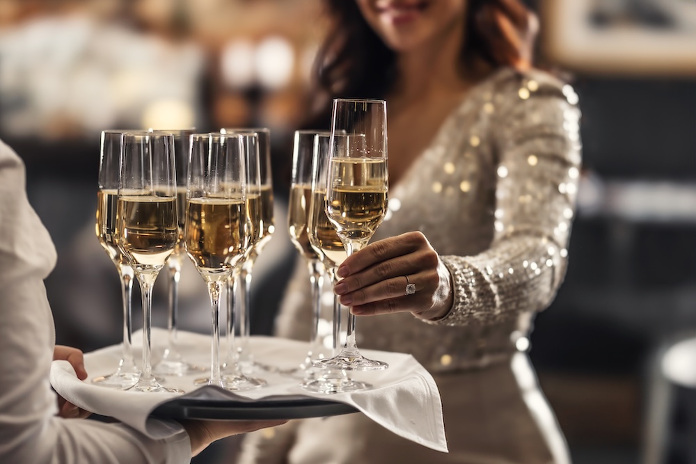 A bartender holds a plate with pink champagne and a woman in a cocktail dress takes one of the glasses at an event, party or wedding.
