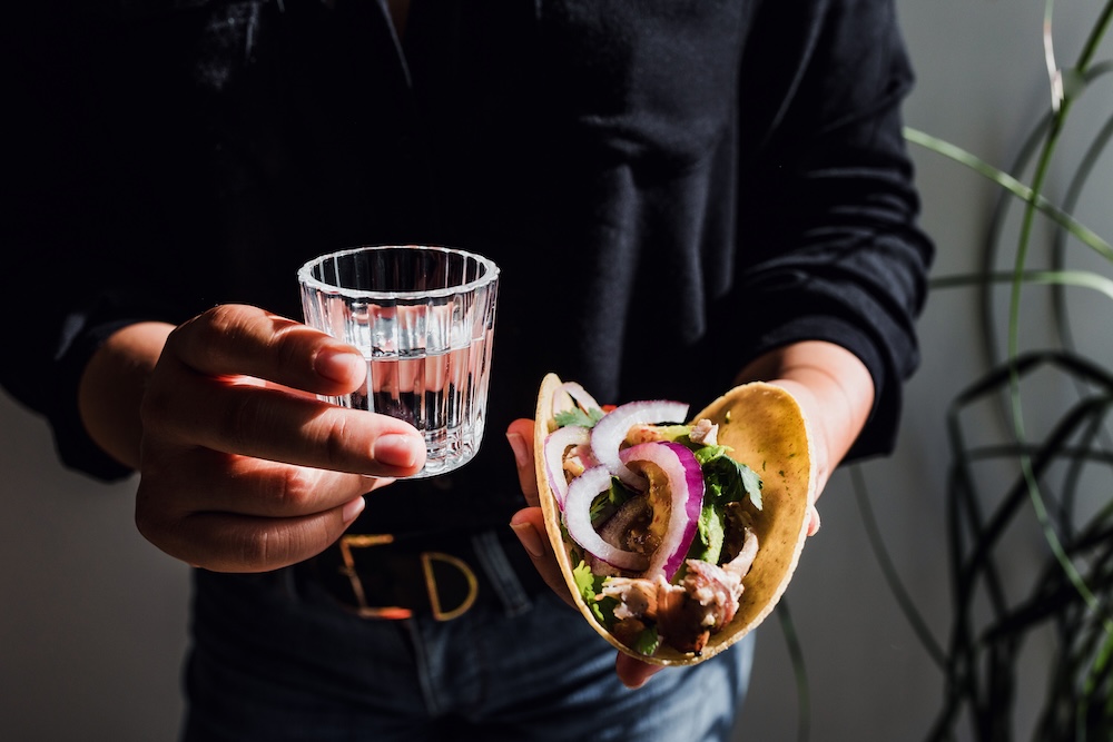 Two hands holding a taco and a mezcal shot. 